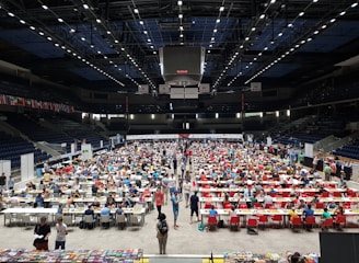 An indoor sports arena filled with rows of tables, each occupied by people engaged in competitive activities. The ceiling is high, with numerous lights illuminating the large space. The audience seating on either side is empty, and there are flags hanging from the upper level.