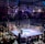 A crowded indoor arena with a boxing ring at the center, surrounded by an audience seated at tables. The scene is illuminated by overhead lights, casting a vibrant glow over the ring where two fighters are preparing for a match. Spectators are engaged, capturing the event with cameras and enjoying the lively atmosphere.