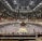 A large indoor arena packed with spectators seated in tiers around a central platform. The platform features traditional Japanese decor, indicating it may be used for sumo wrestling. The atmosphere is bustling, with people filling the seats and many spectators visible in the foreground.