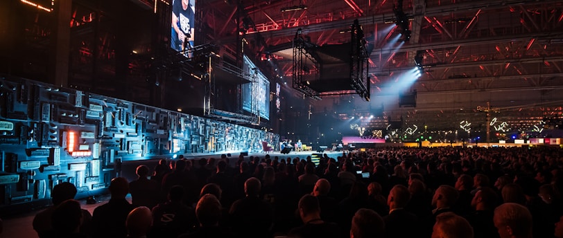 A large indoor event with a stage featuring bright screens and spotlights, attended by a substantial crowd. The venue has a high ceiling with industrial beams and red lighting accents. The atmosphere resembles a concert or a conference with a focus on visual effects.
